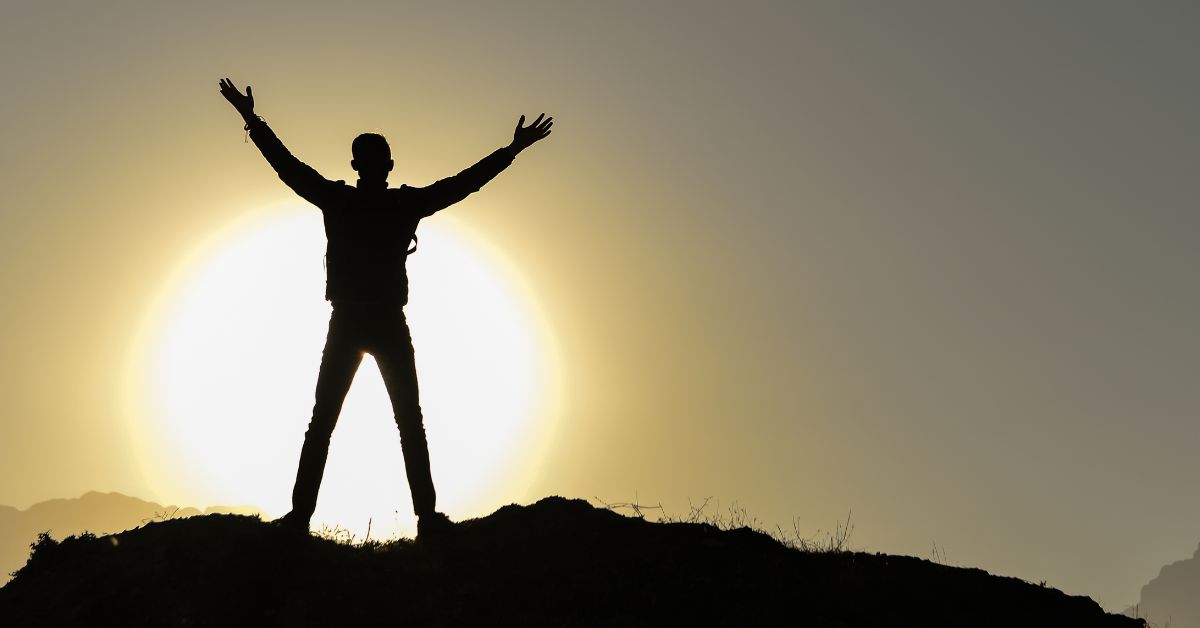 A hiker in a large, green field, backlit by a bright sun setting behind a distant mountain range, legs striding with a proud determination